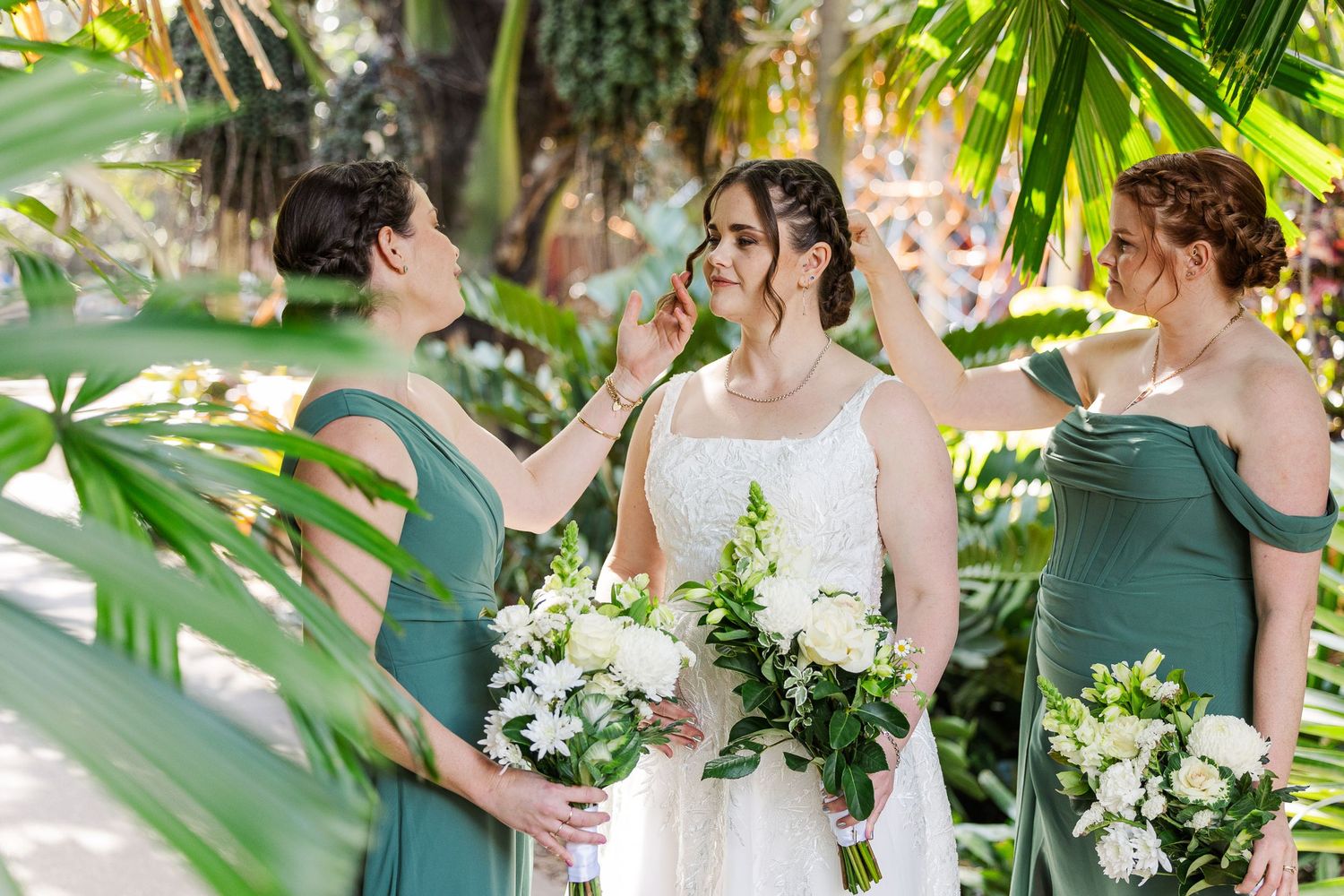 Bride in white gown with bridesmaids in sage green dresses holding white floral bouquets outdoors.