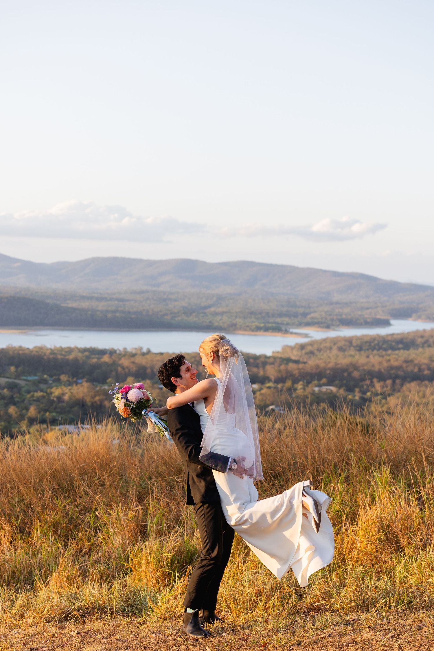 Groom lifting bride in white dress and veil on scenic hilltop with lake and mountains in background.