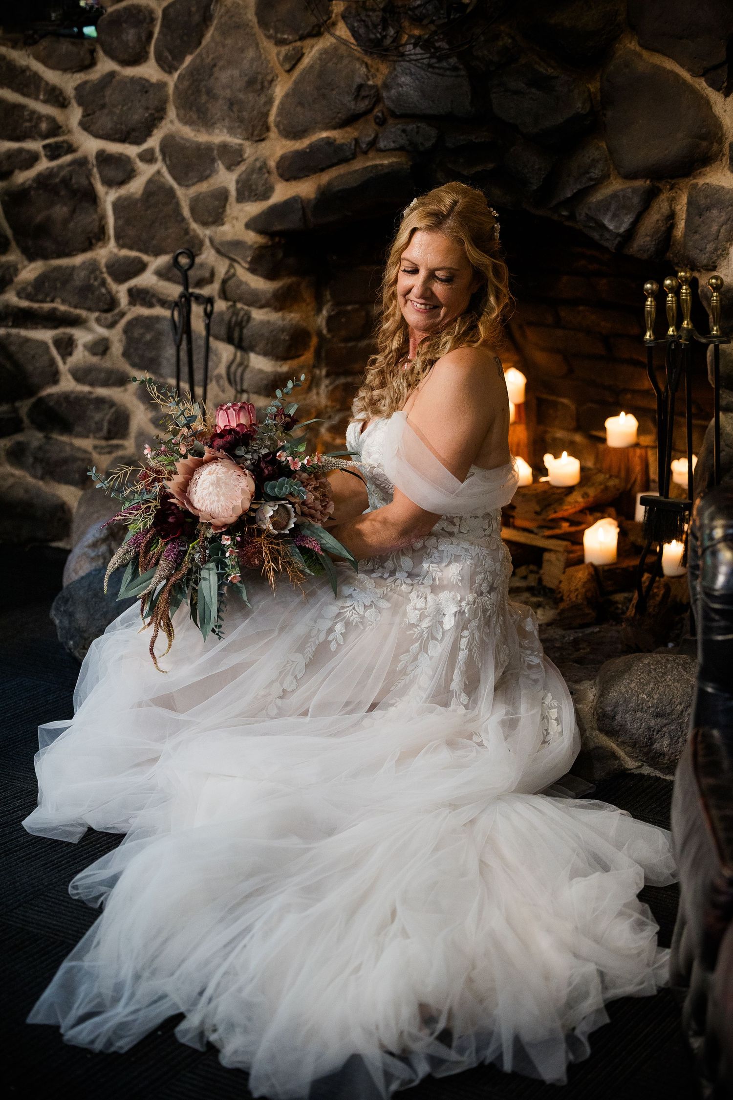 Smiling bride in lace tulle gown holds pink bouquet by candlelit stone fireplace.