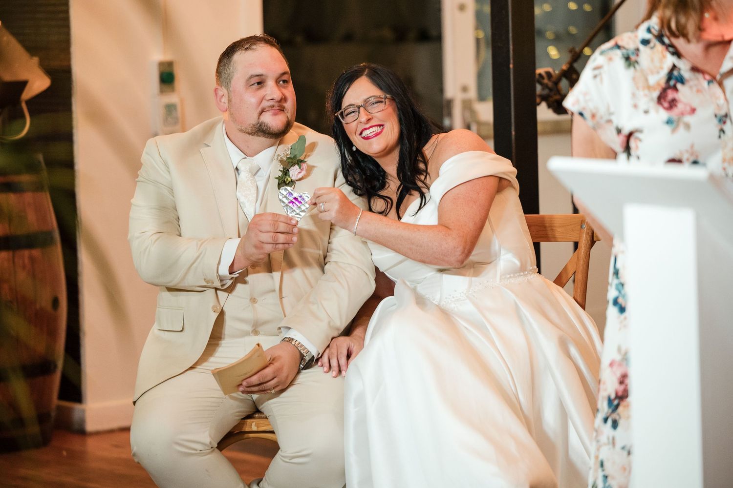 Bride and groom laughing joyfully during wedding reception toasts, bride in white gown, groom in tan suit with boutonniere.