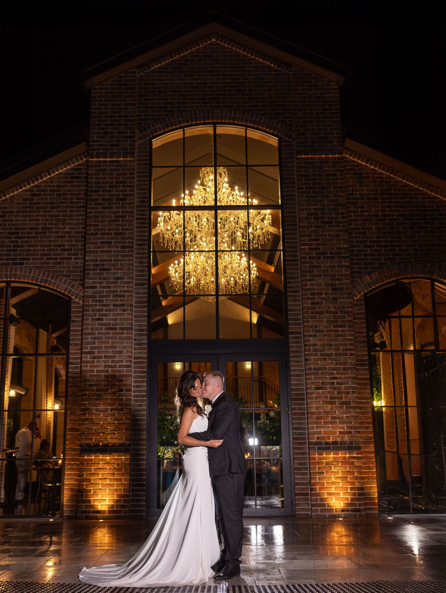 Elegant wedding couple embraces under glowing chandelier in front of brick venue with dramatic lighting at night.