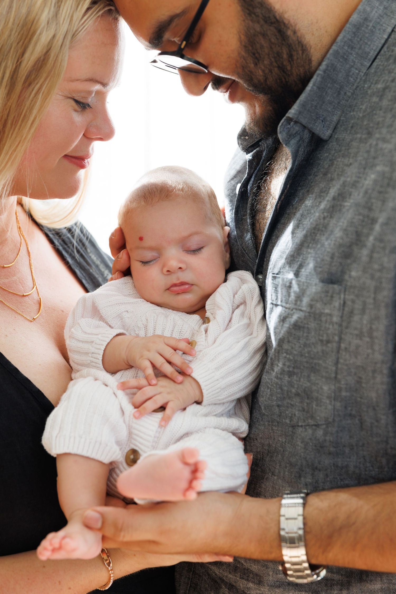 A sleeping baby in a white outfit is cradled lovingly between two adults in an intimate family portrait.