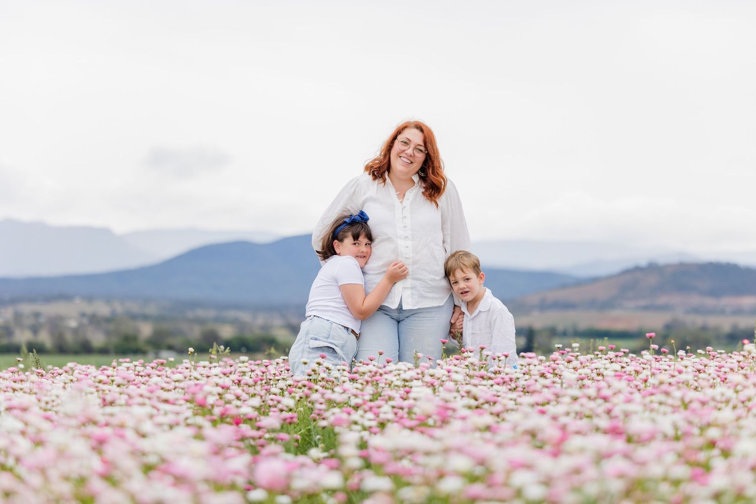 A mother and two children smile in a pink flower field with mountain views.