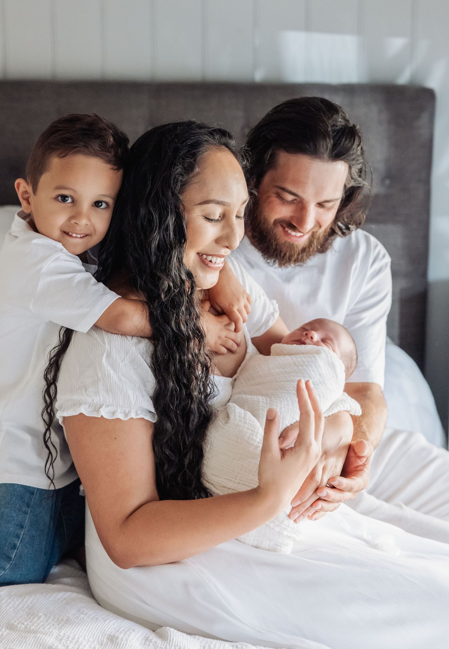 A family lies together on white bedding in a series of intimate lifestyle portraits showing tender moments.