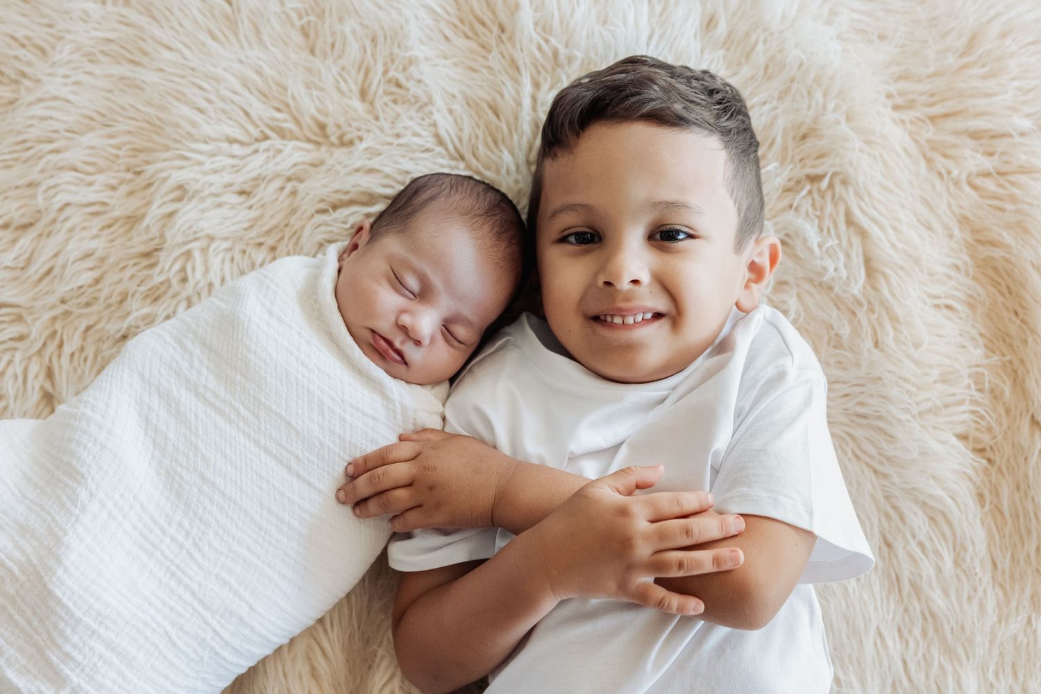 Two children in white clothing lying together on a soft white fur blanket.