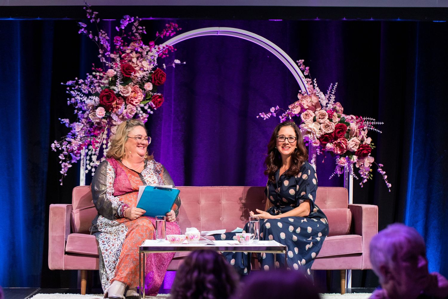 Two people sit on a pink couch under an arch decorated with pink and purple flowers during a stage event.
