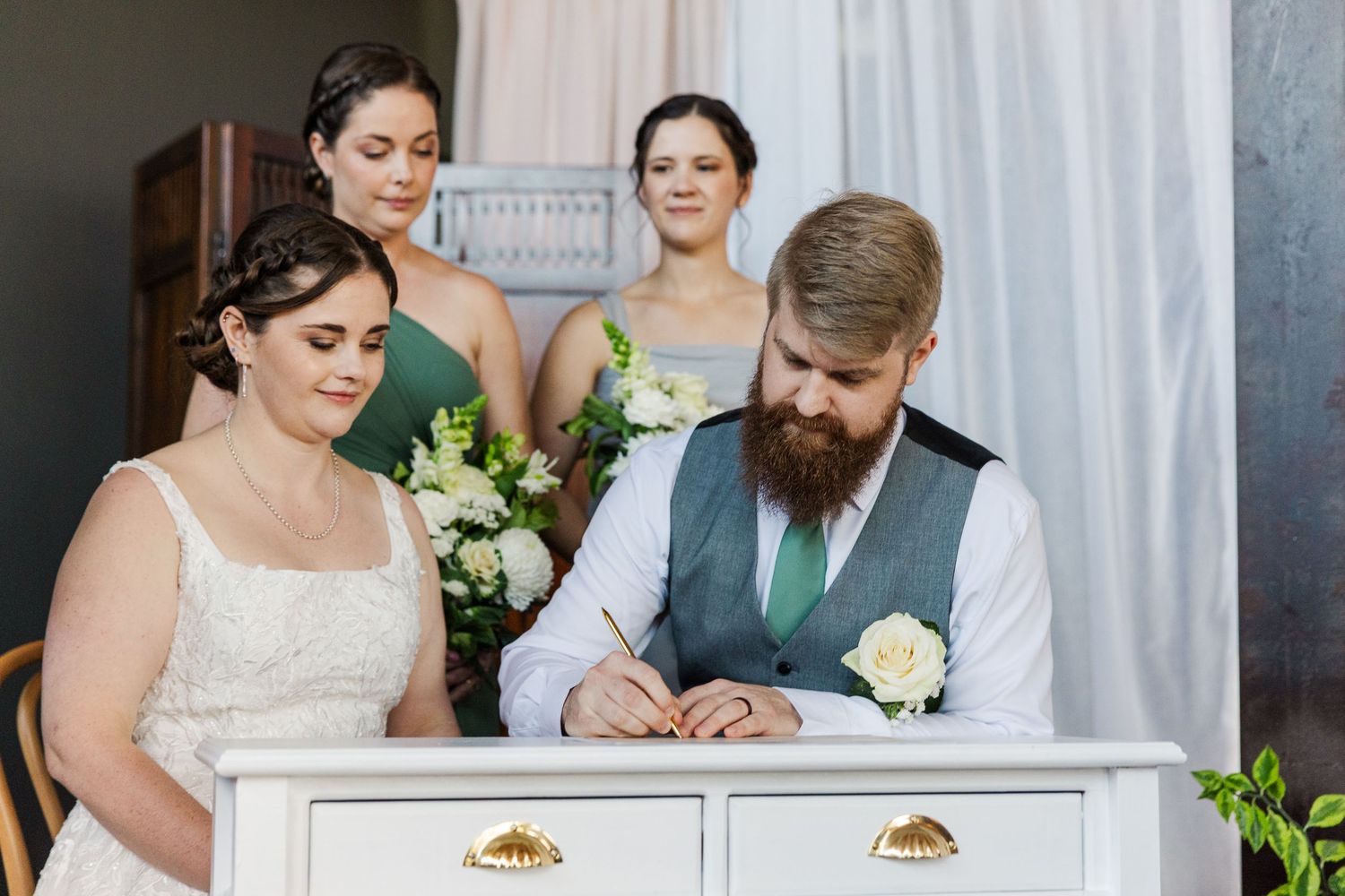 Bride and groom signing wedding registry at white desk with two bridesmaids holding bouquets standing behind them.