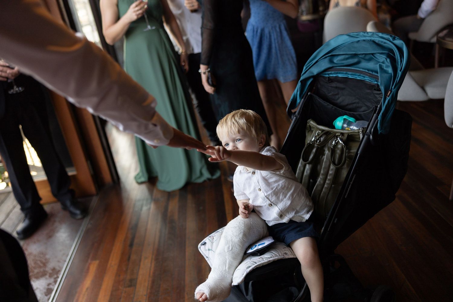A baby in white outfit sits in a stroller reaching out to an adult at a wedding reception.