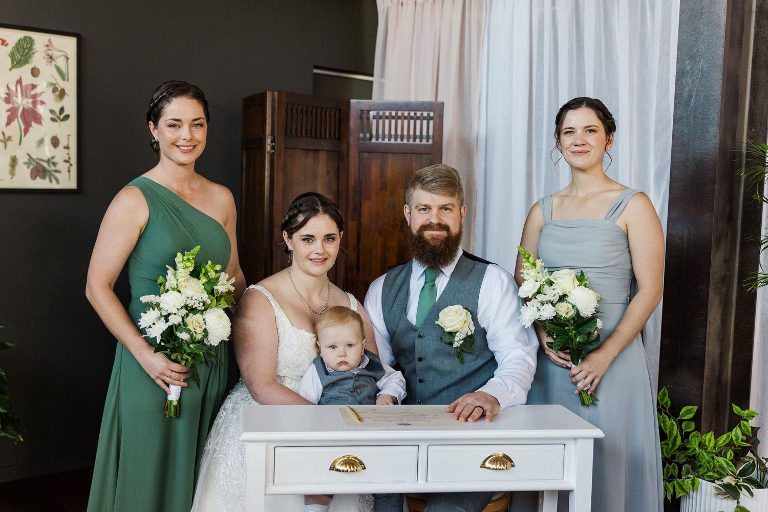 Bride and groom with baby pose with two bridesmaids in green and gray dresses holding white bouquets at elegant wedding.