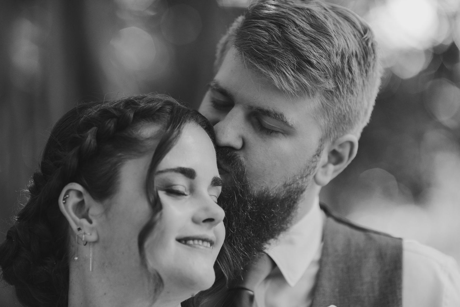 Black and white wedding photo of a bearded groom kissing his bride on the cheek as she smiles softly.