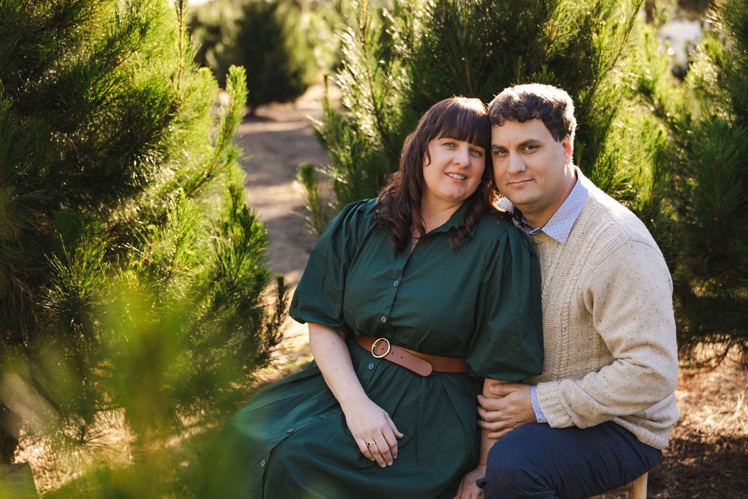 A couple in casual attire shares intimate moments in a sunlit outdoor setting with evergreen trees in the background.