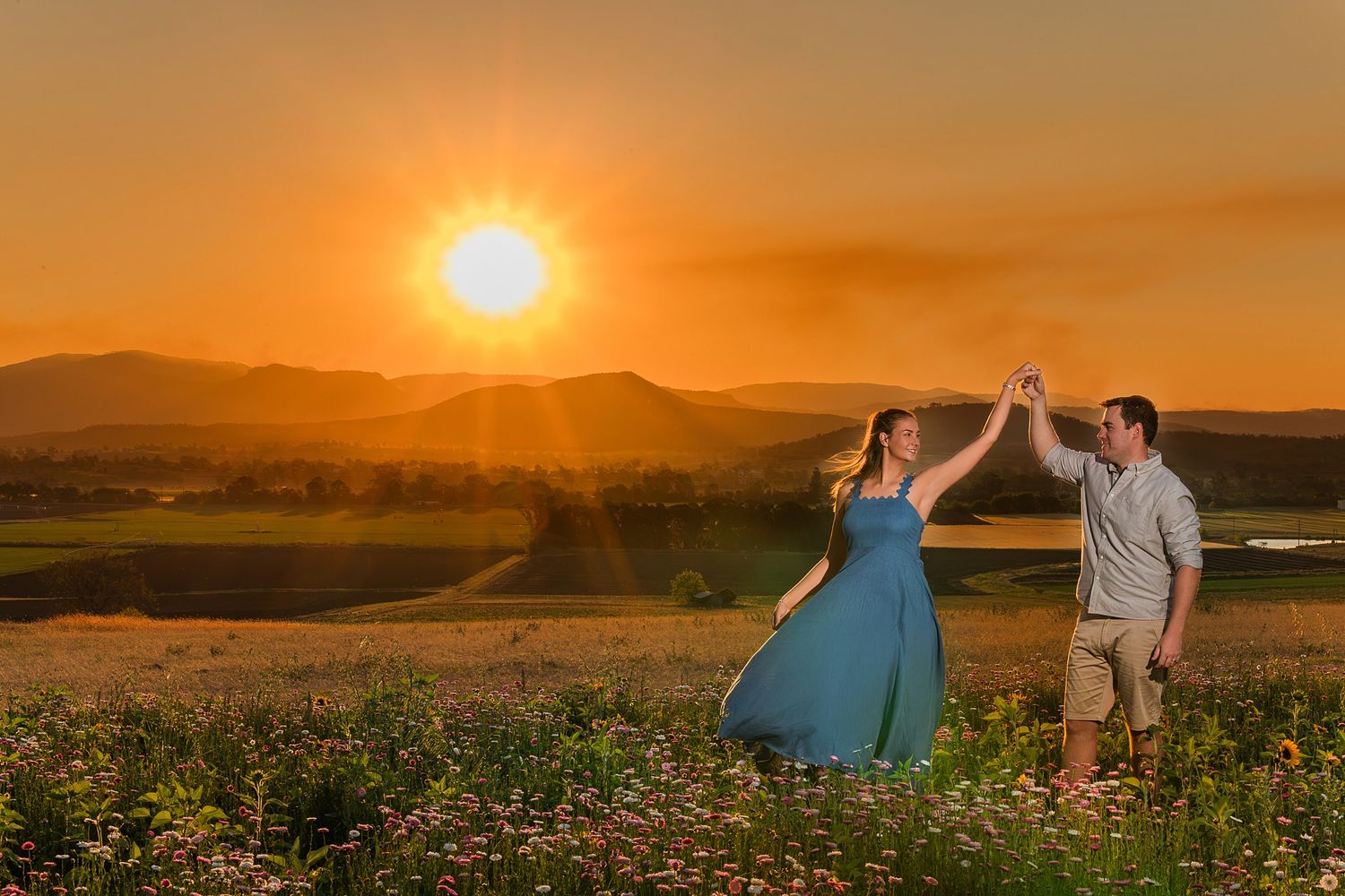 Couple holding hands and dancing in a wildflower meadow at golden sunset, with rolling mountains and glowing orange sky behind them.