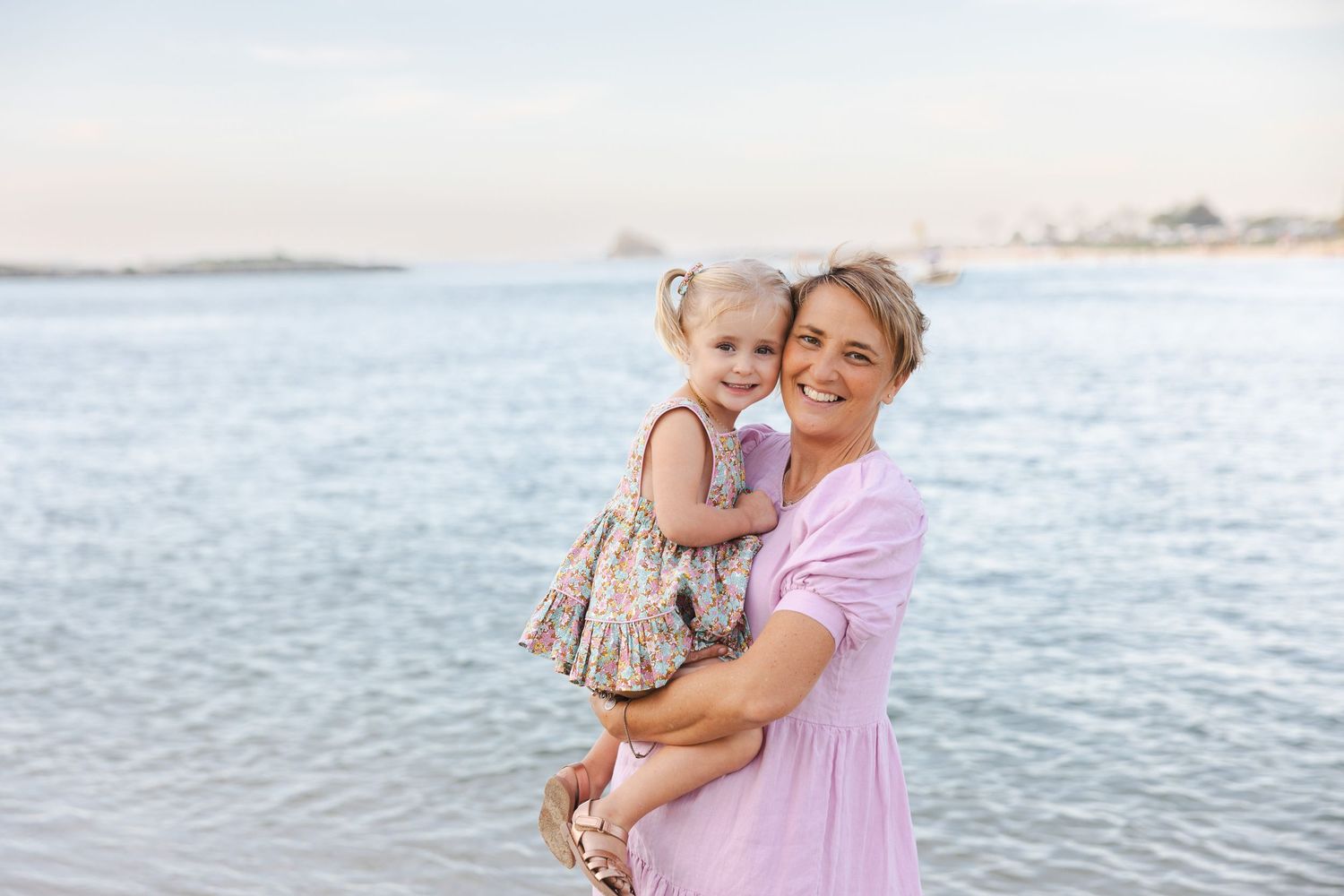 A smiling woman in a pink dress holds a young girl in a floral dress on a sunny beach by the water.