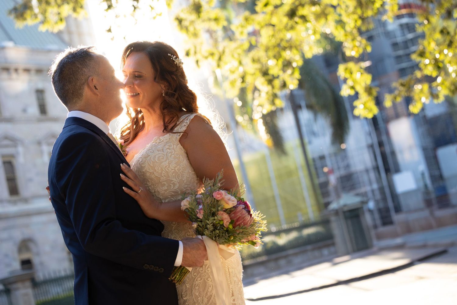 A romantic wedding couple shares a tender moment outdoors with sunlit trees in the background.