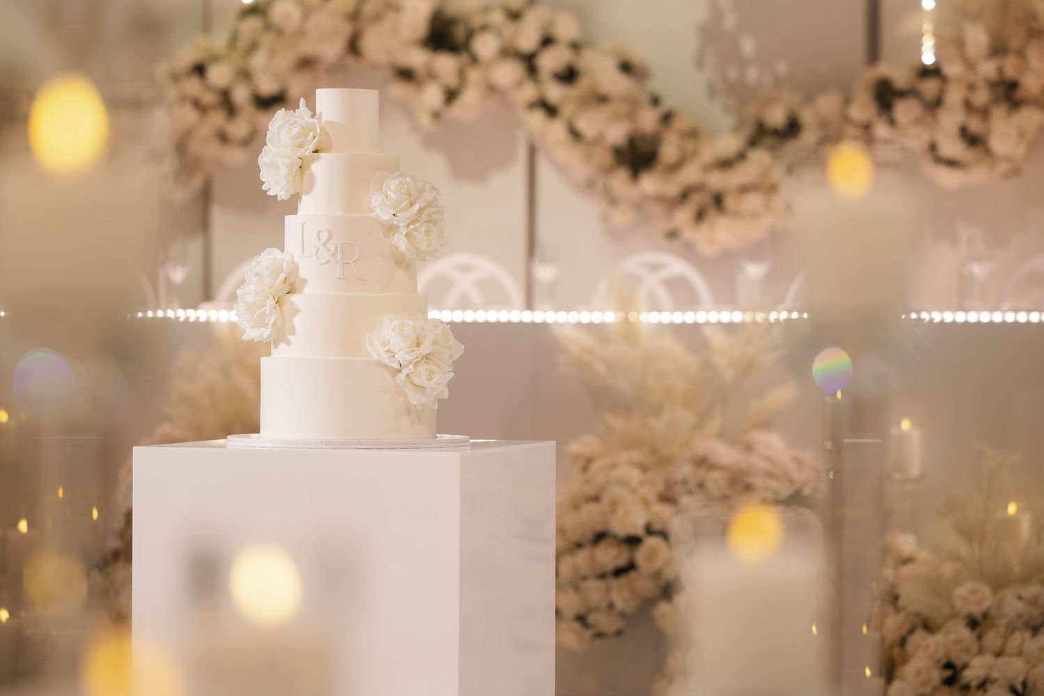 White wedding cake with multiple tiers decorated with fresh flowers and illuminated by soft glowing lights in the background.