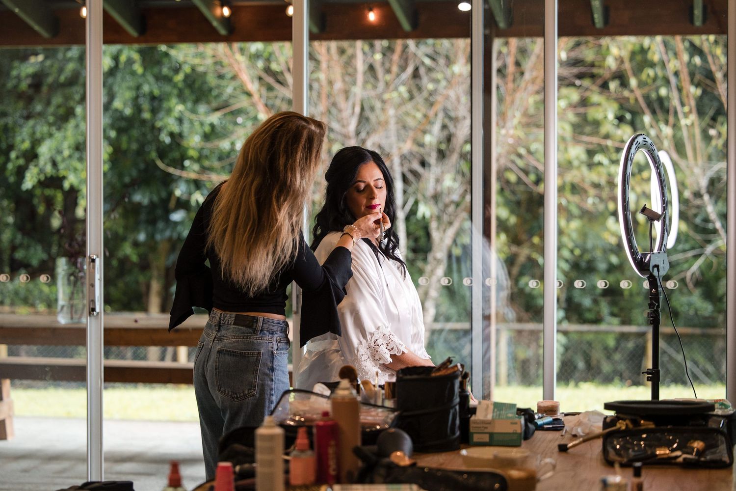 Makeup artist applying cosmetics to a woman in a bright studio with ring light and lush greenery backdrop.