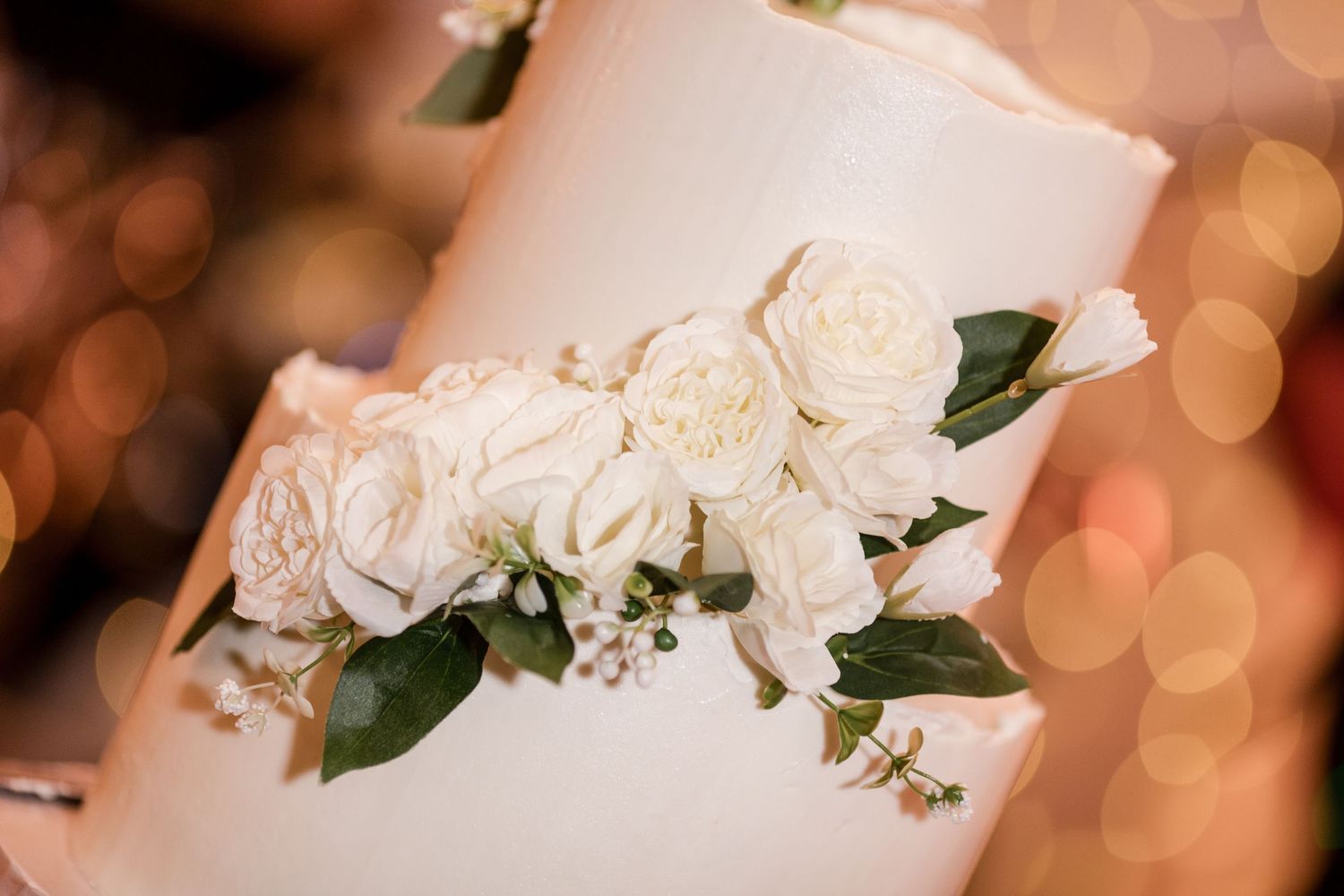 Elegant white wedding cake adorned with fresh white roses and green leaves, with soft bokeh lights in the background.