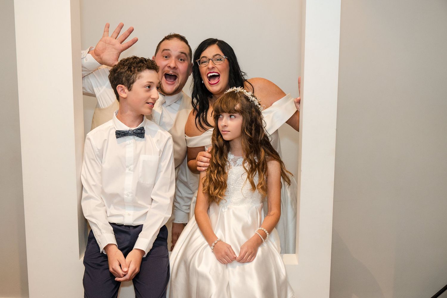 Family of four posing playfully in a photo booth at a wedding, kids dressed formally, parents laughing and waving.