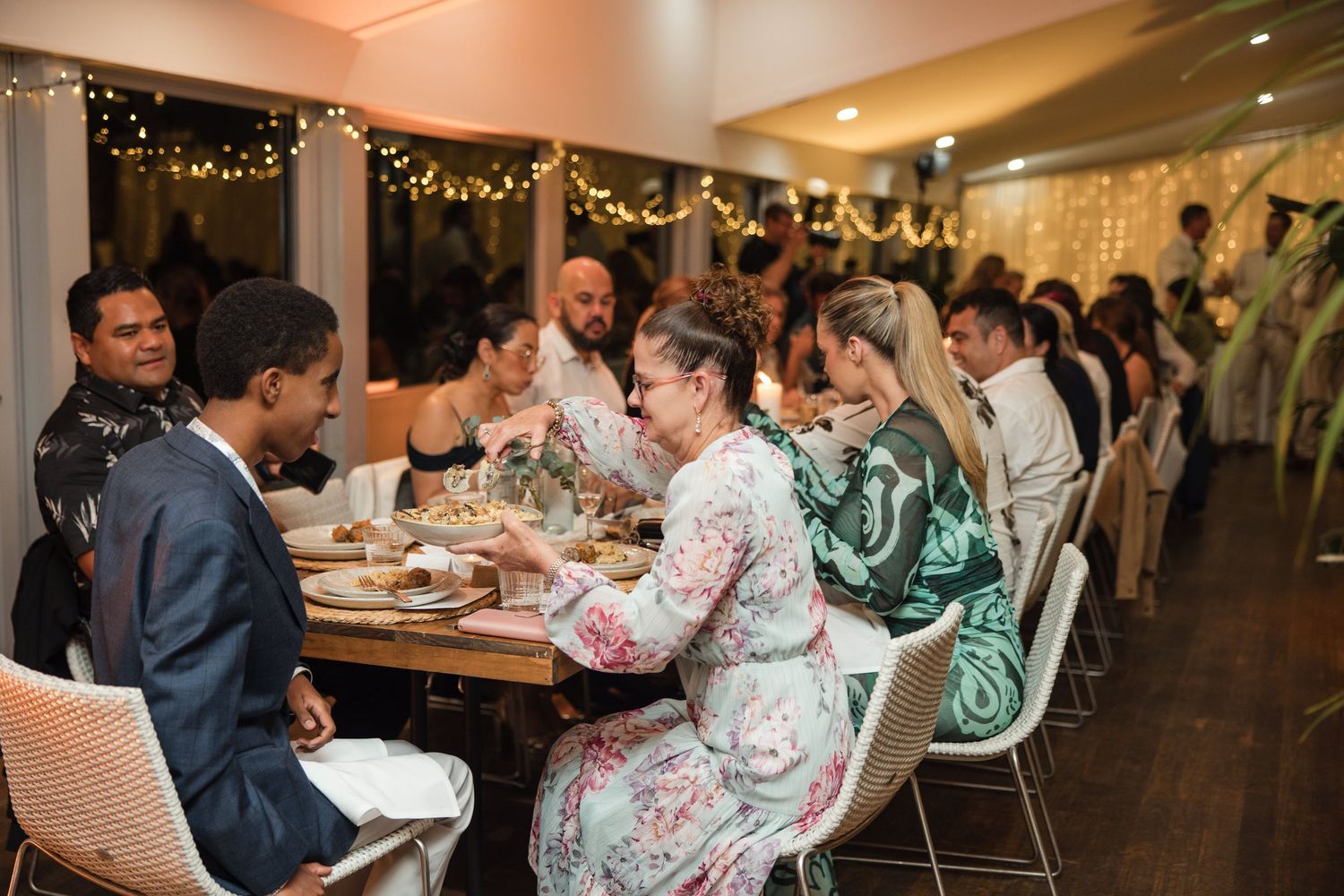 Guests enjoying a festive dinner party at a long table decorated with warm string lights indoors.