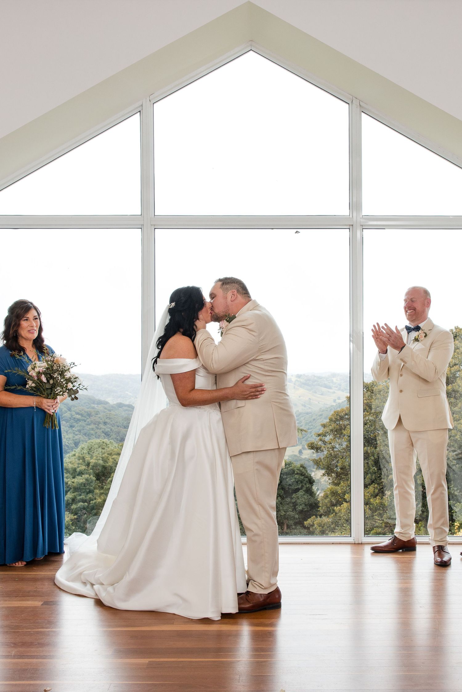 Bride and groom share first kiss at wedding altar with panoramic forest views through chapel windows.