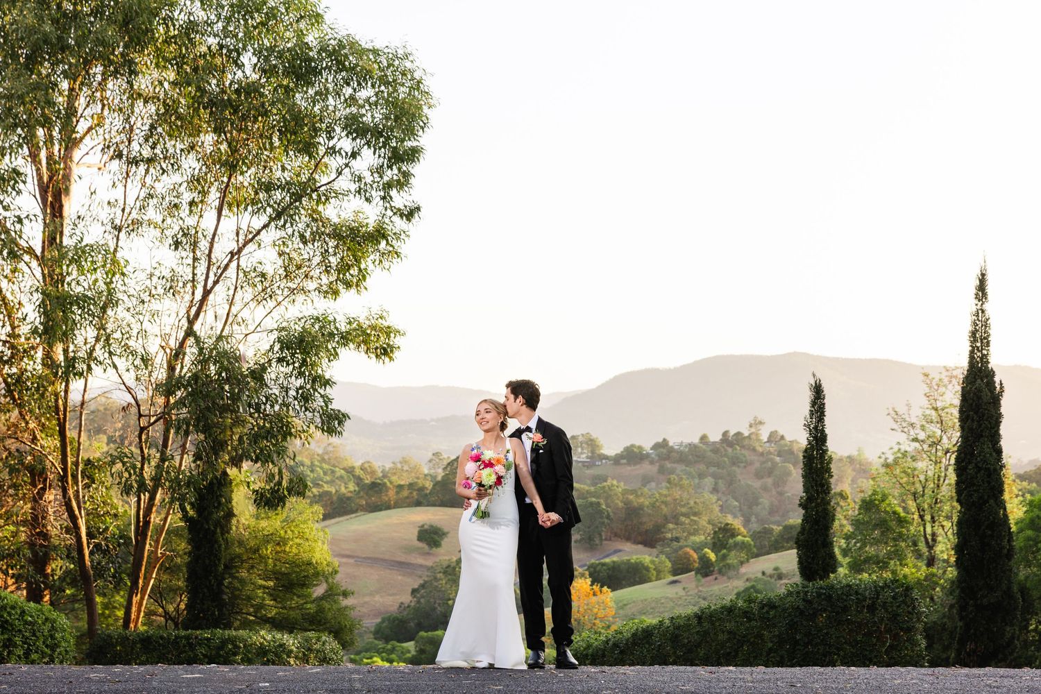 Couple embraces in a romantic sunset moment on a hilltop with cypress trees and golden light.
