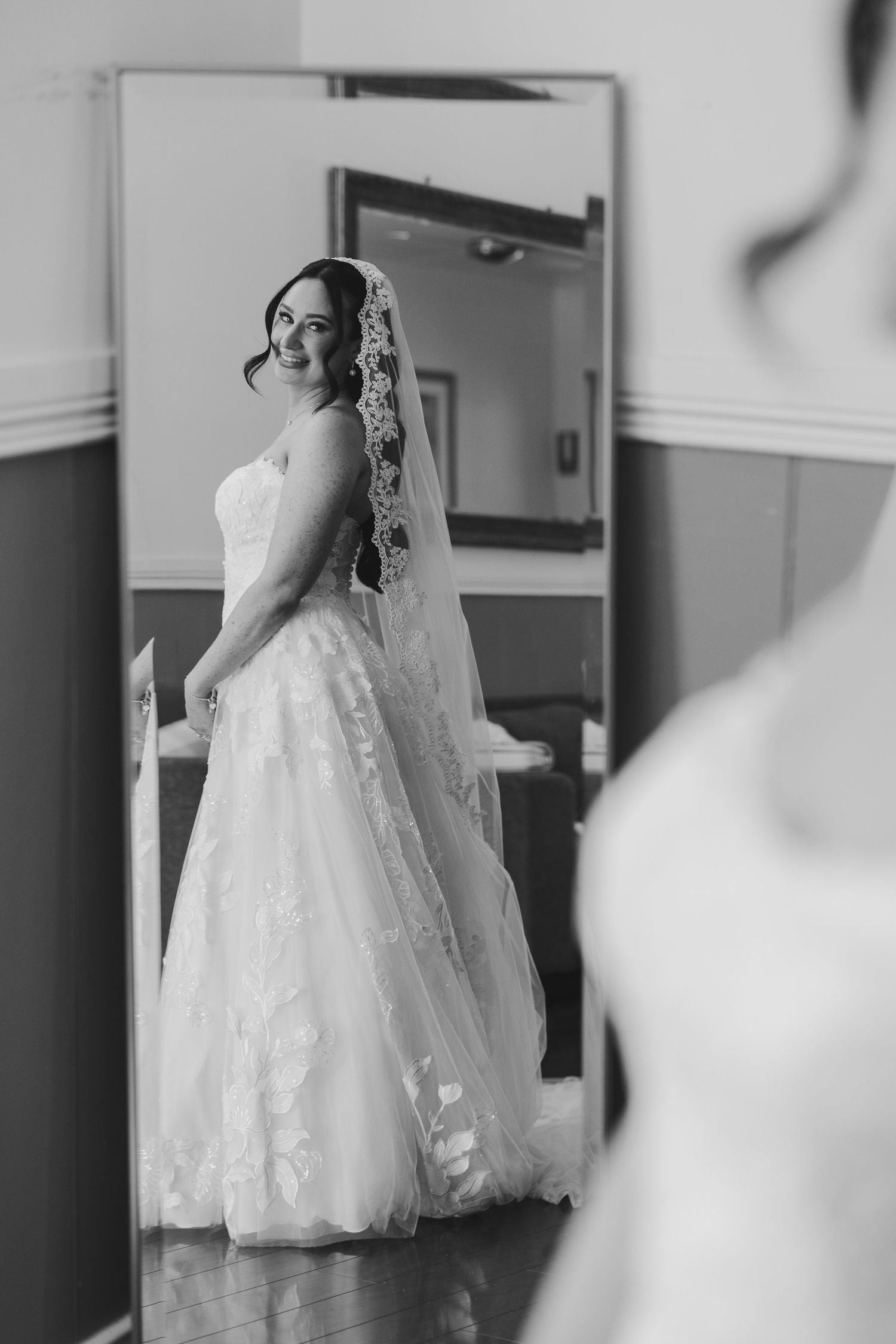 Bride in lace wedding gown and veil smiling at her reflection in a full-length mirror in black and white.