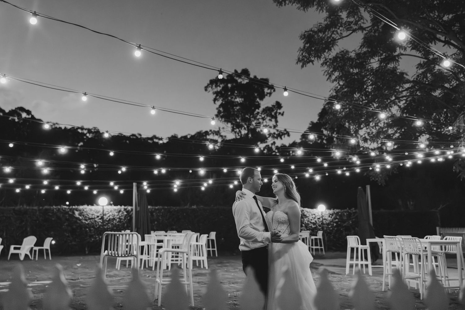 A couple shares a romantic moment under string lights at an outdoor evening reception with empty chairs in the background.