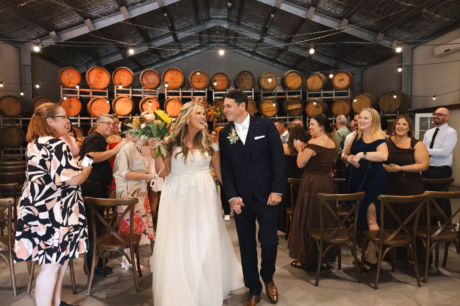 Bride and groom walk down the aisle in a rustic winery wedding venue with oak barrels and cheering guests.
