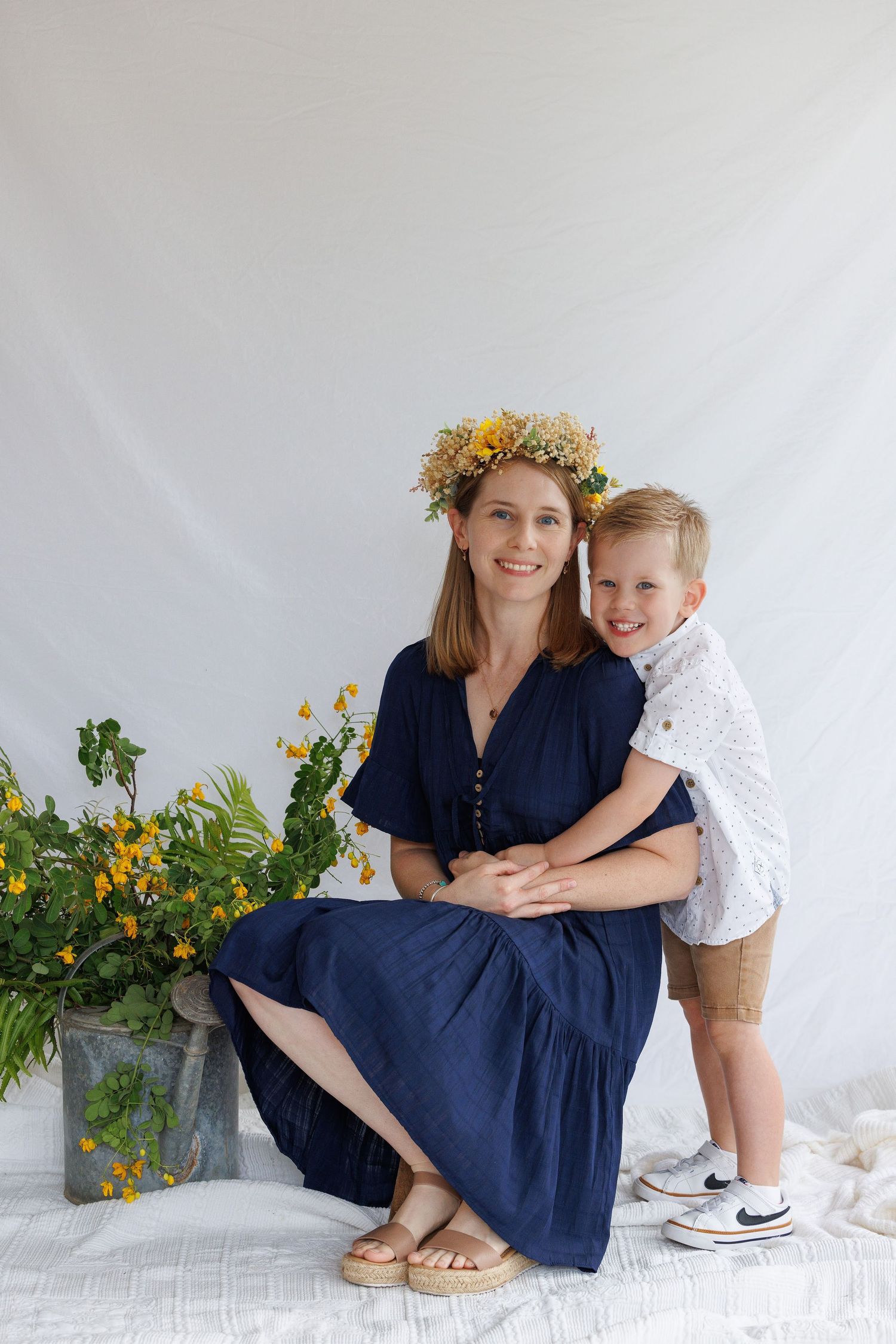 A pair shares tender moments while sitting on white floor with yellow flowers and greenery in the background.