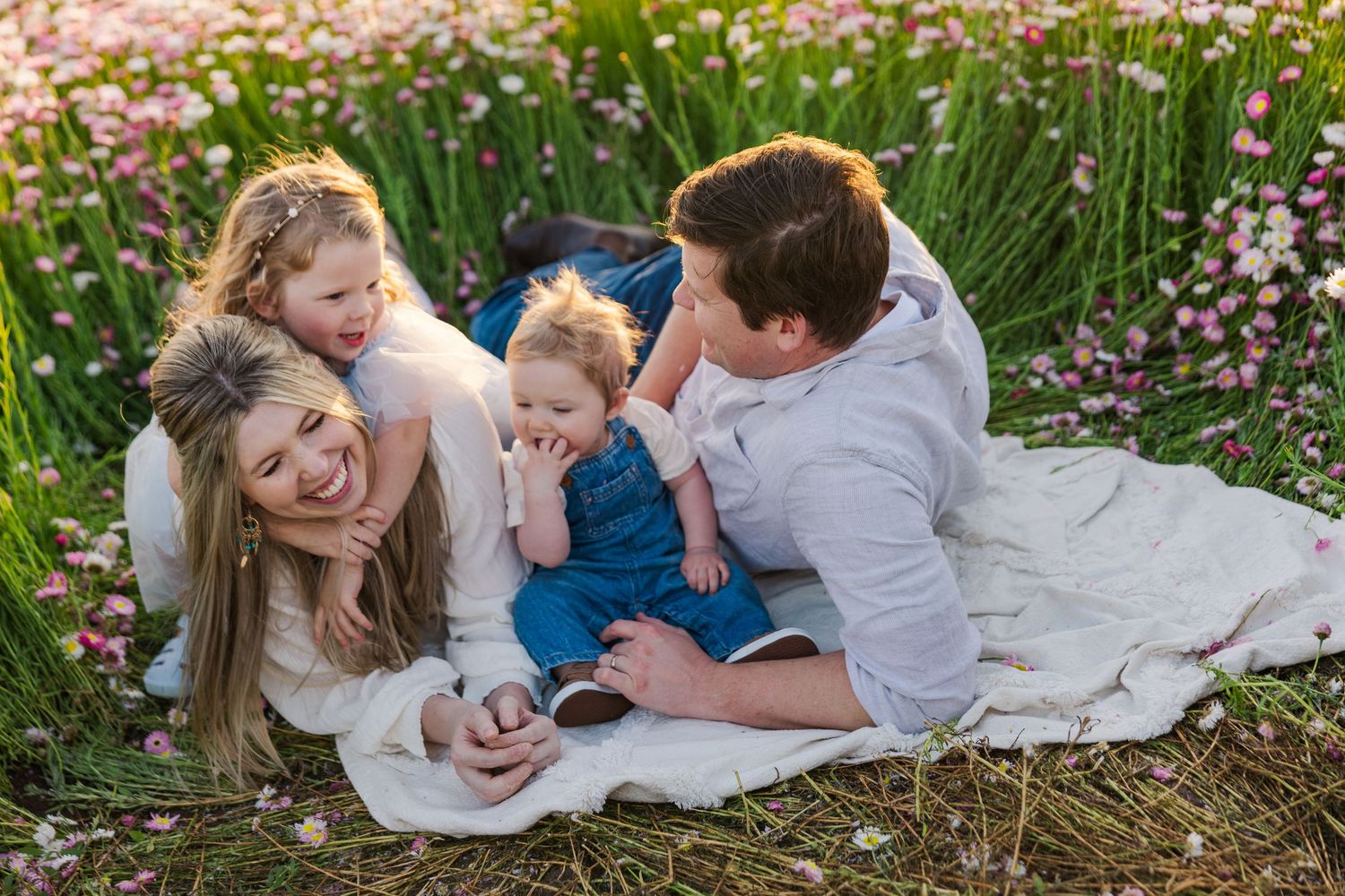 A family lounges together on a white blanket surrounded by pink wildflowers and tall grass in a garden setting.