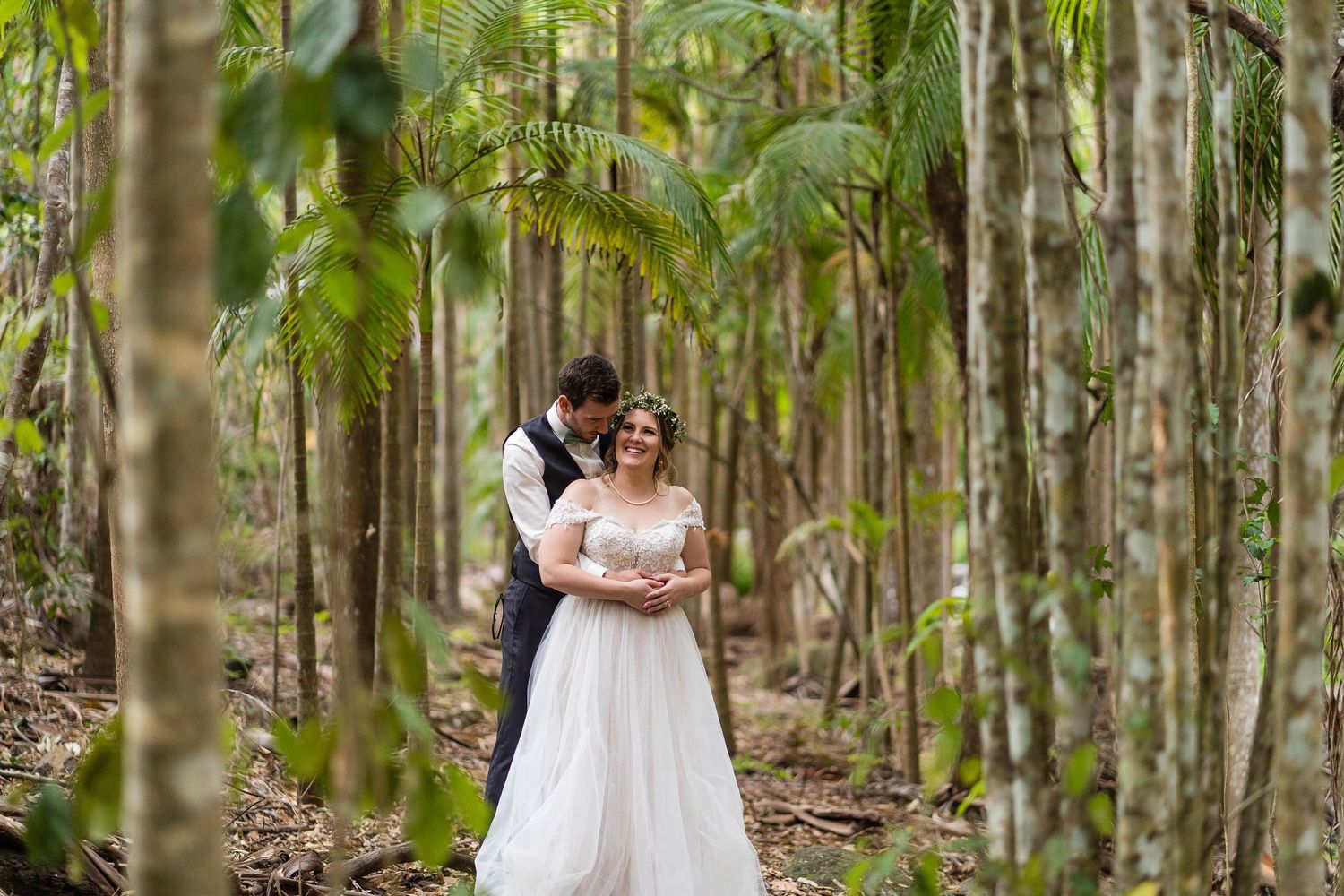 A couple in wedding attire stands together in a lush forest setting with tall trees and green foliage.