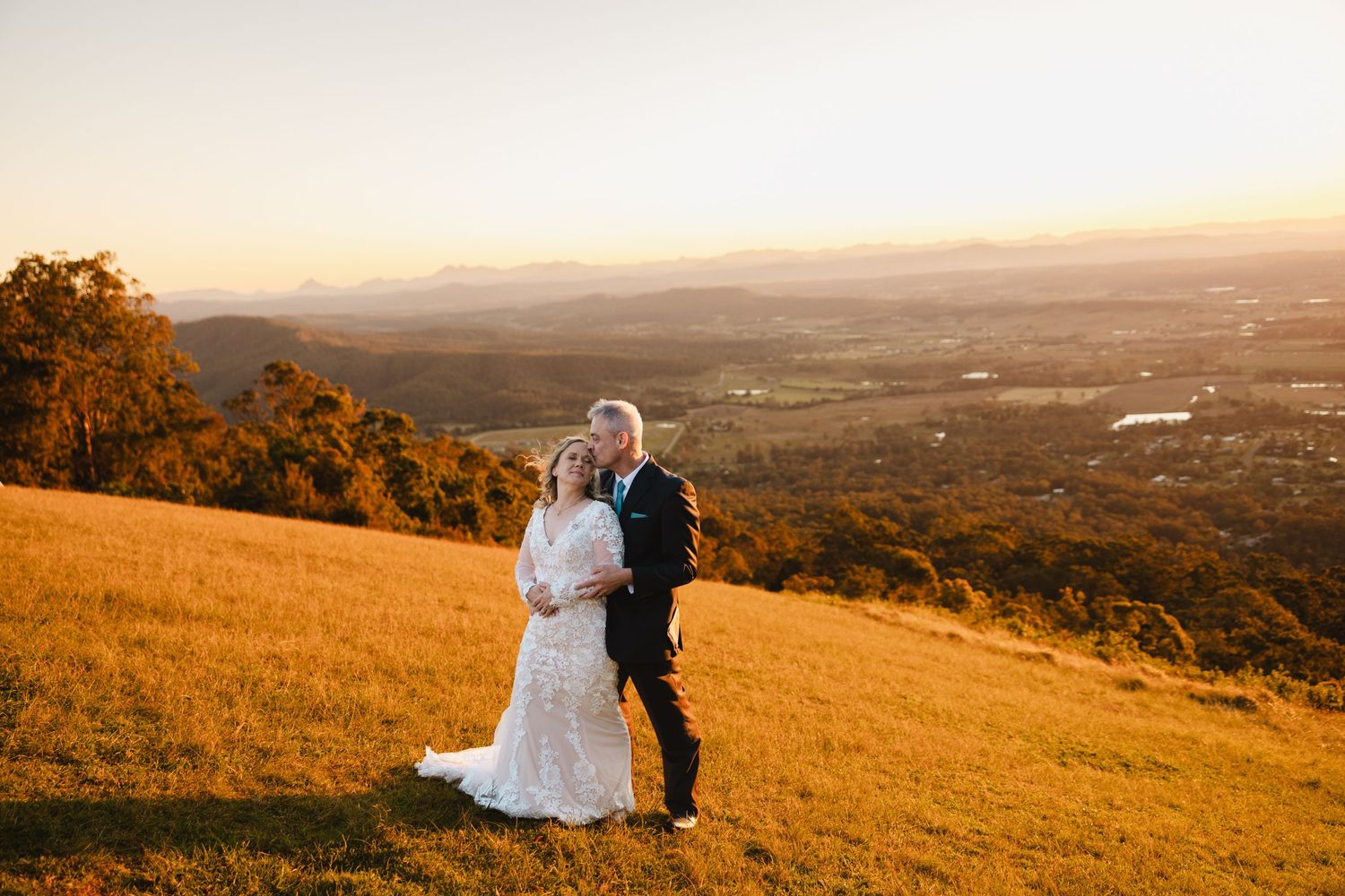 A couple in wedding attire stands on a golden hillside at sunset overlooking a scenic valley landscape.
