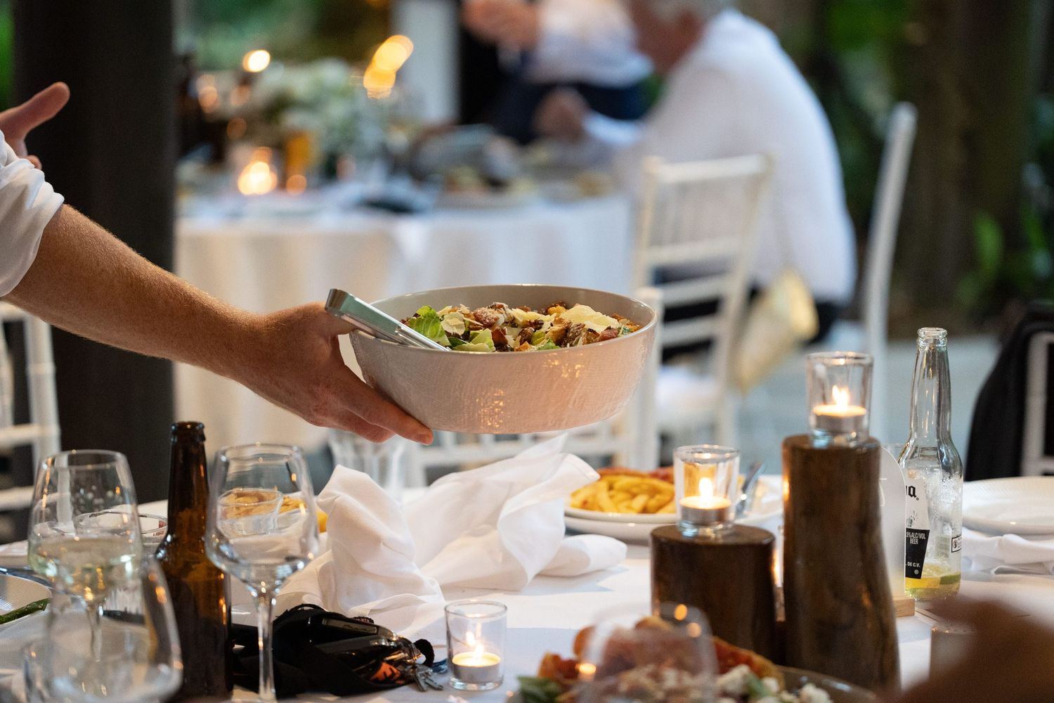 A bowl of food being passed across an elegant dinner table set with glassware and candles during a special occasion.