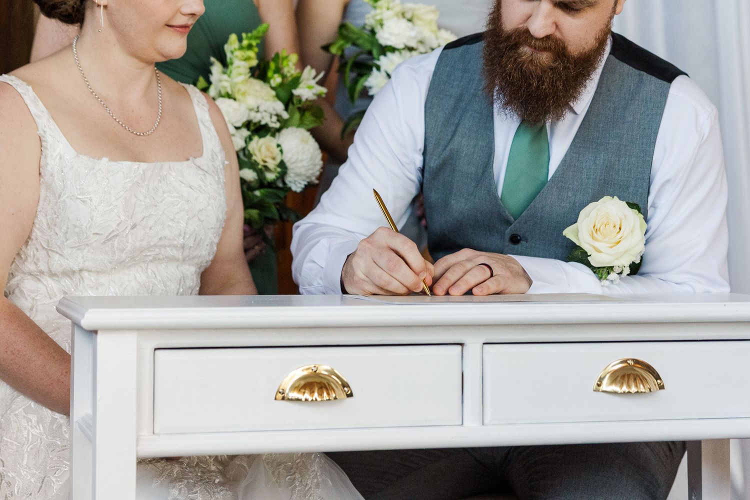 Bride and bearded groom signing wedding certificate at white table with gold handles during ceremony.