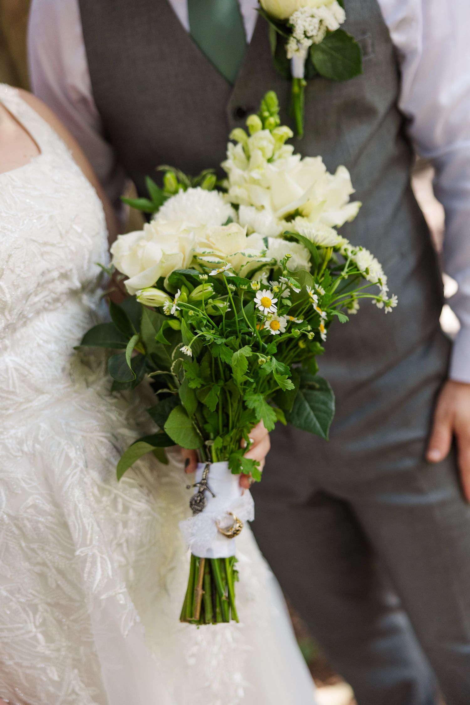 Bride holding a lush white wedding bouquet with greenery, standing beside groom in gray vest.