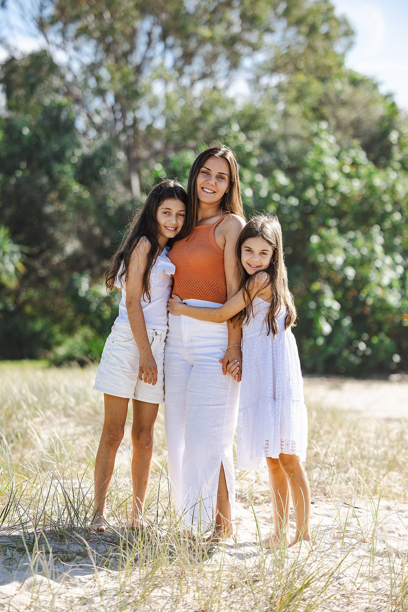 Three figures in coordinating white and orange summer outfits pose together on a sunny beach with natural vegetation.