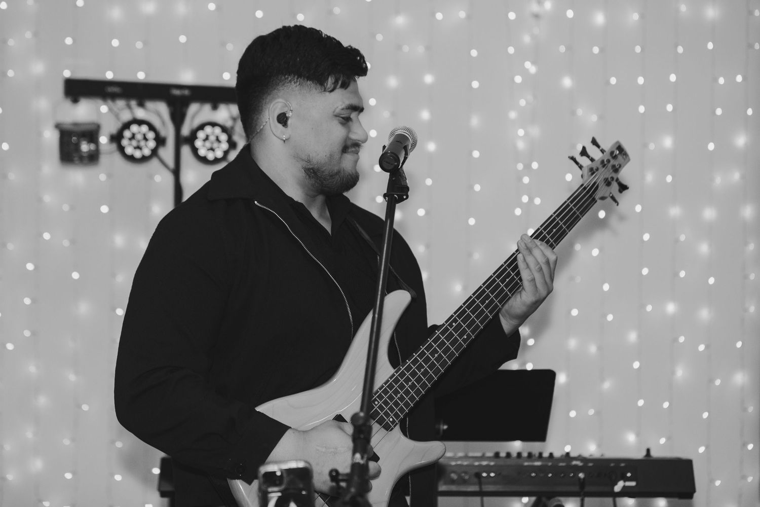 Black and white photo of a male musician playing bass guitar and singing into a microphone on stage.