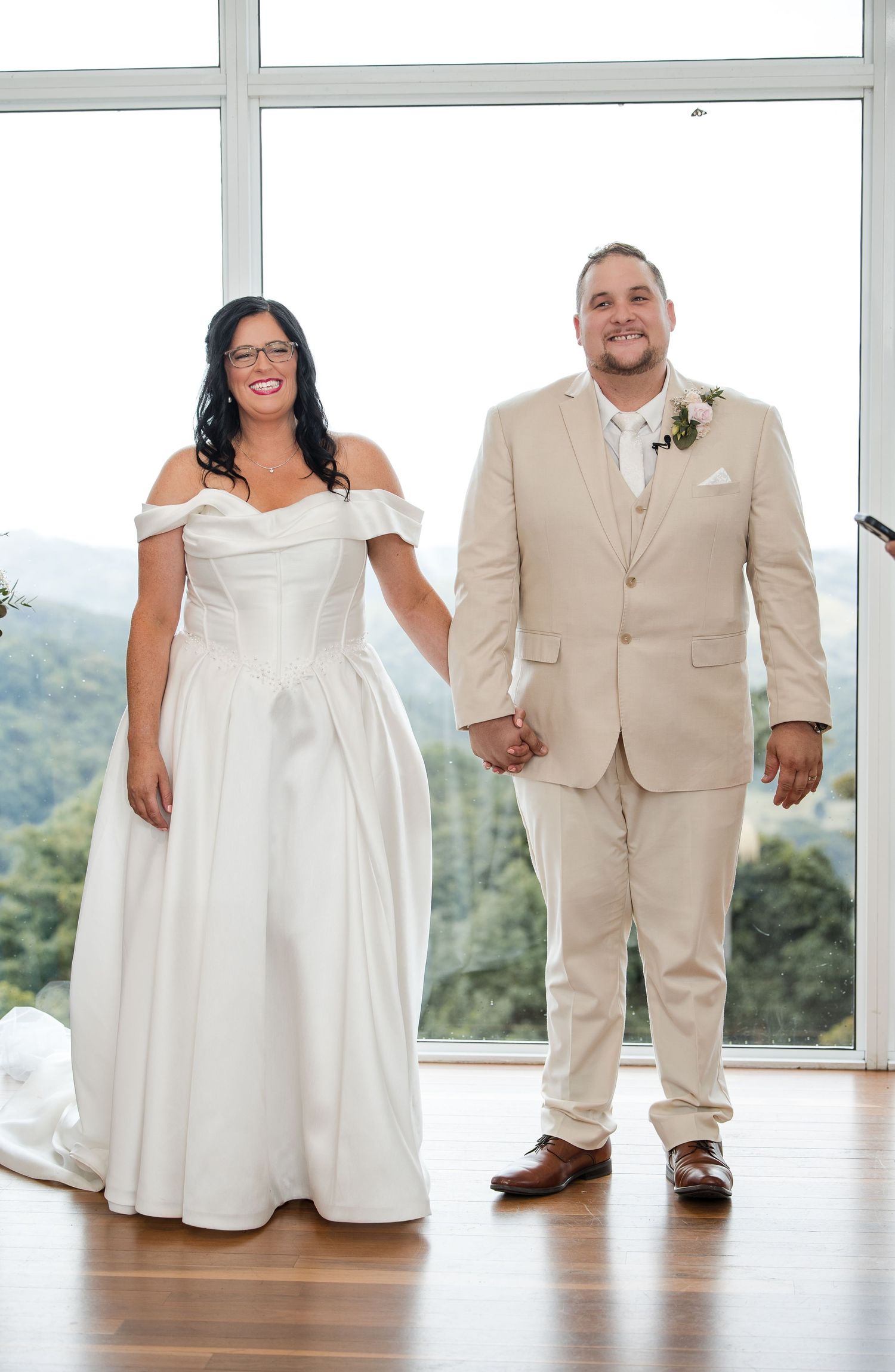 A bride in a white off-shoulder gown and groom in a beige suit hold hands at their wedding ceremony with mountain views.