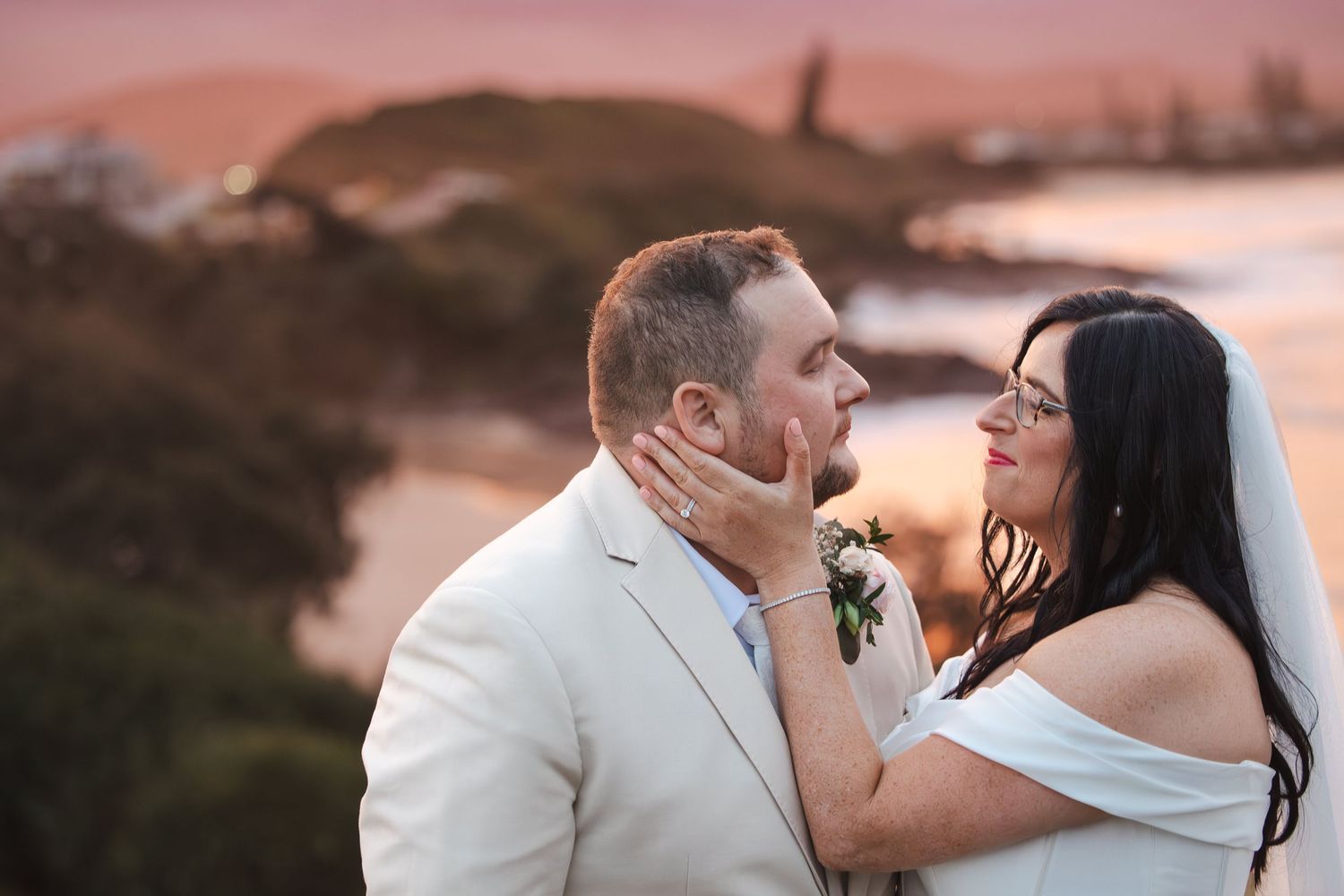 Bride and groom share a romantic sunset moment on a coastal cliff, bride in white gown touching grooms face tenderly.