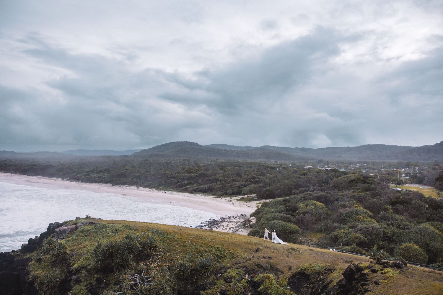 Aerial view of a coastal landscape with lush greenery, a sandy beach, and dramatic cloudy skies over rolling hills.
