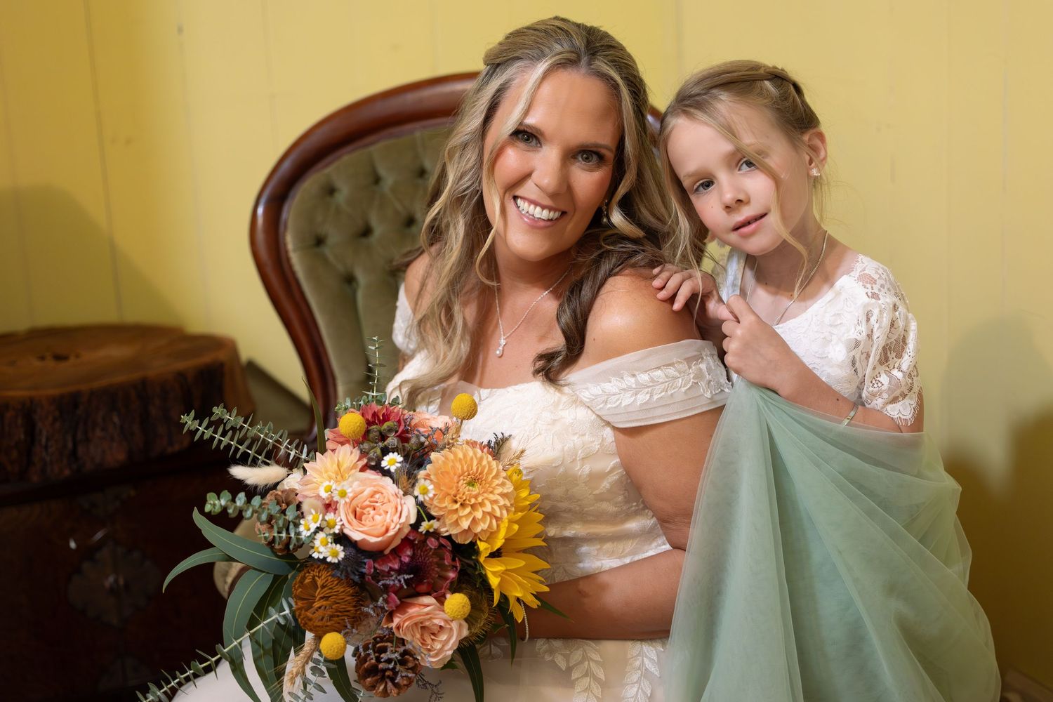 Bride in lace gown holding colorful bouquet poses with young flower girl in white dress at wedding.
