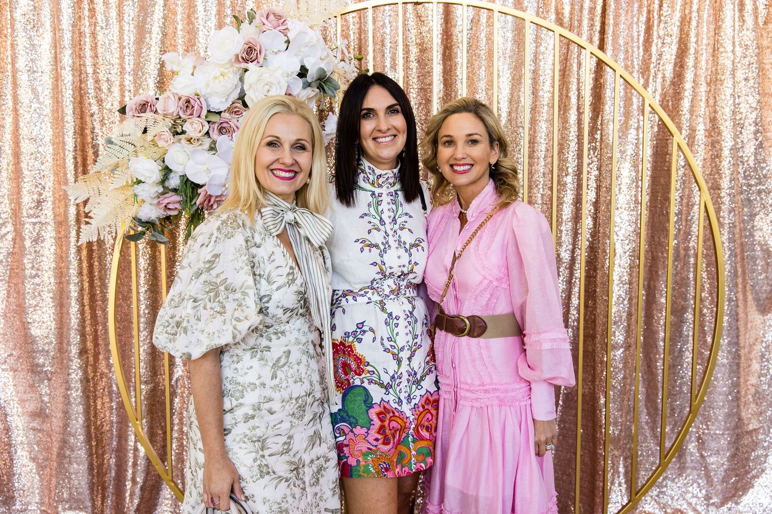 Three women pose in front of a rose gold sequin backdrop with floral decorations at a special event.