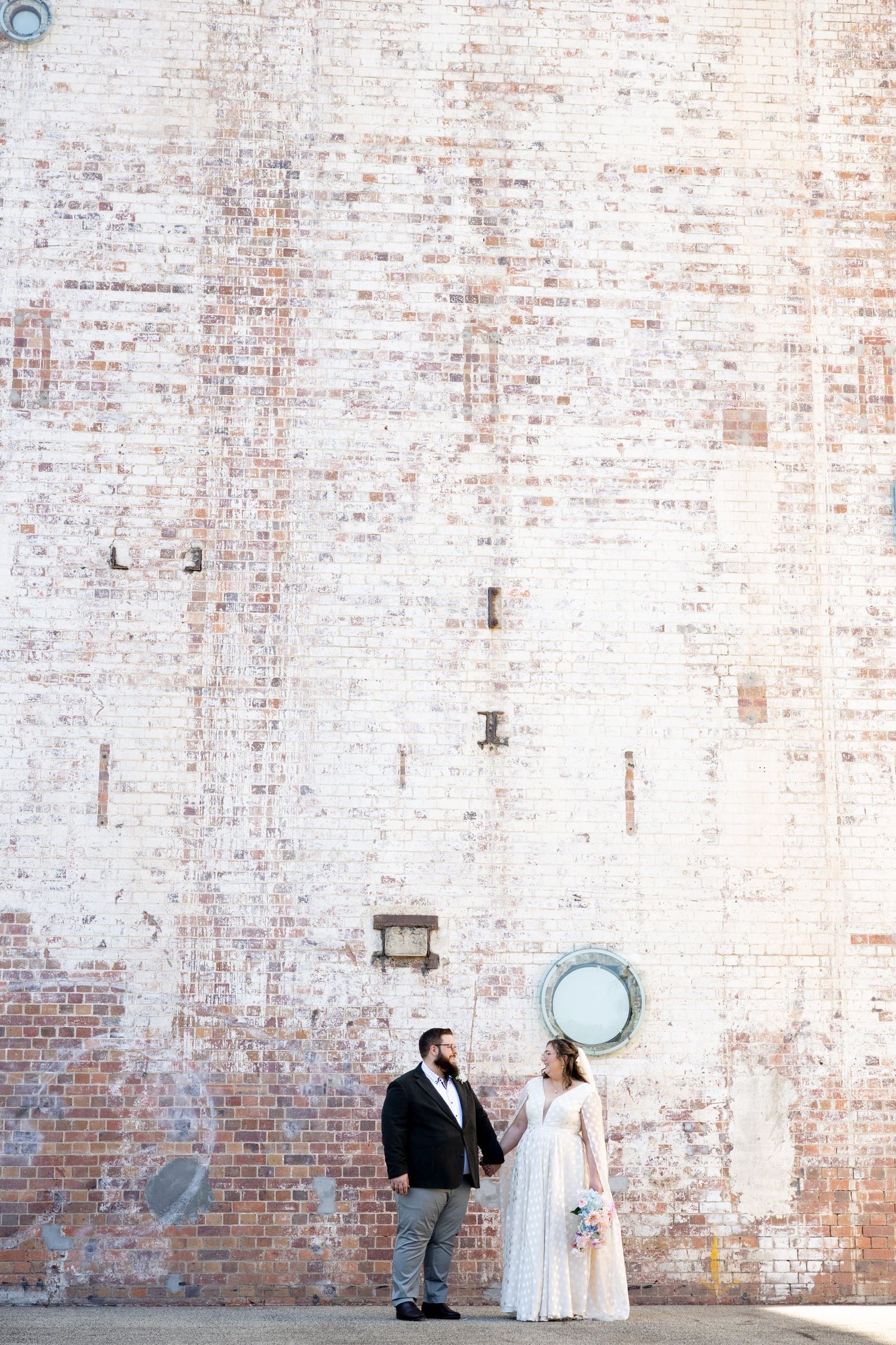 A couple stands together against a weathered brick wall with a circular window, creating an industrial chic wedding portrait.