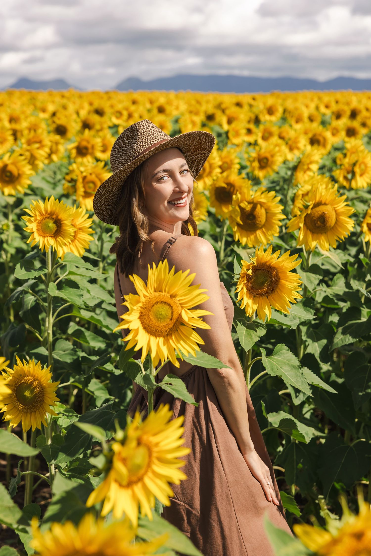 Smiling woman in straw hat and brown dress standing in a vibrant sunflower field under a cloudy sky.