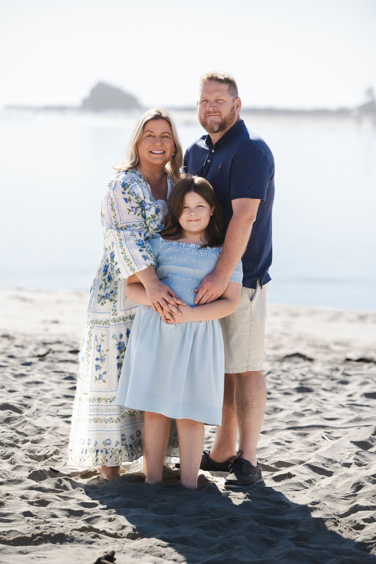 Family of three posing together on a sandy beach, wearing blue and white outfits, smiling with ocean in background.