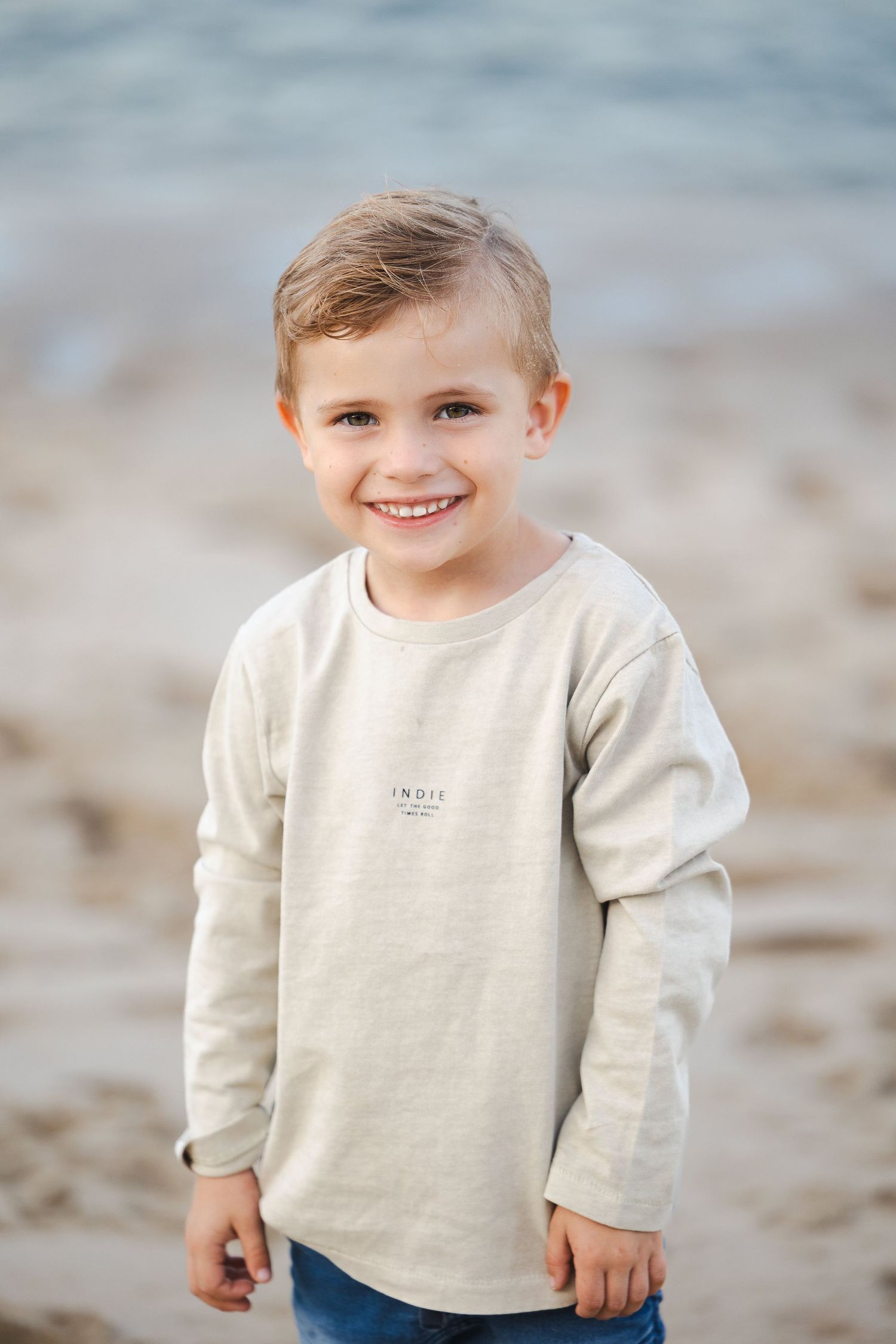 Smiling young boy in a beige long-sleeve shirt standing on a sandy beach with ocean waves in the background.