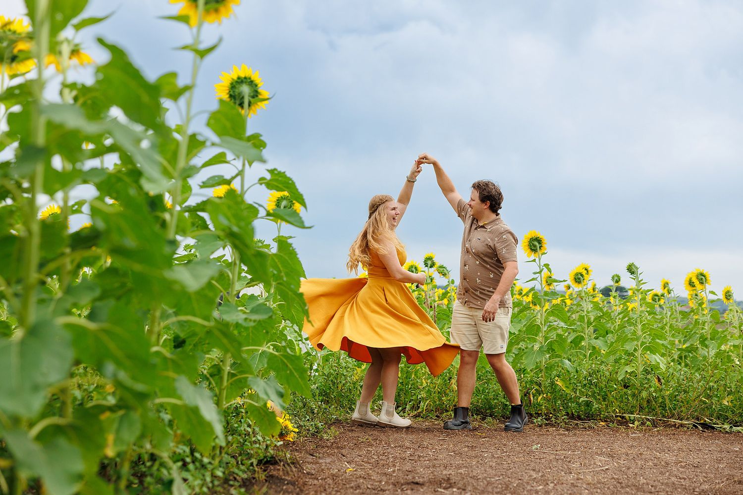 Two people dance together on a dirt path between vibrant sunflower fields on a sunny summer day.