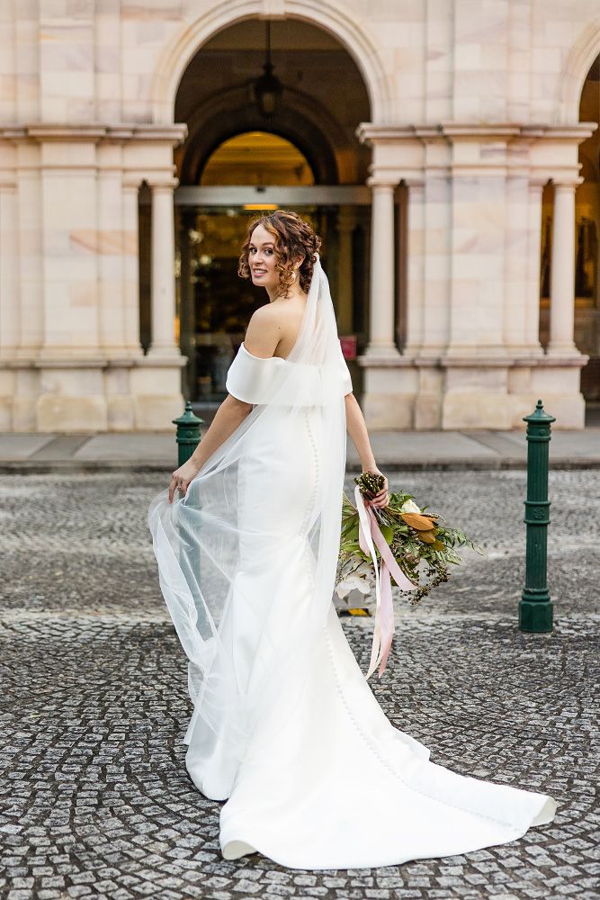 Elegant bride in off-shoulder gown poses by historic archways on cobblestone street.