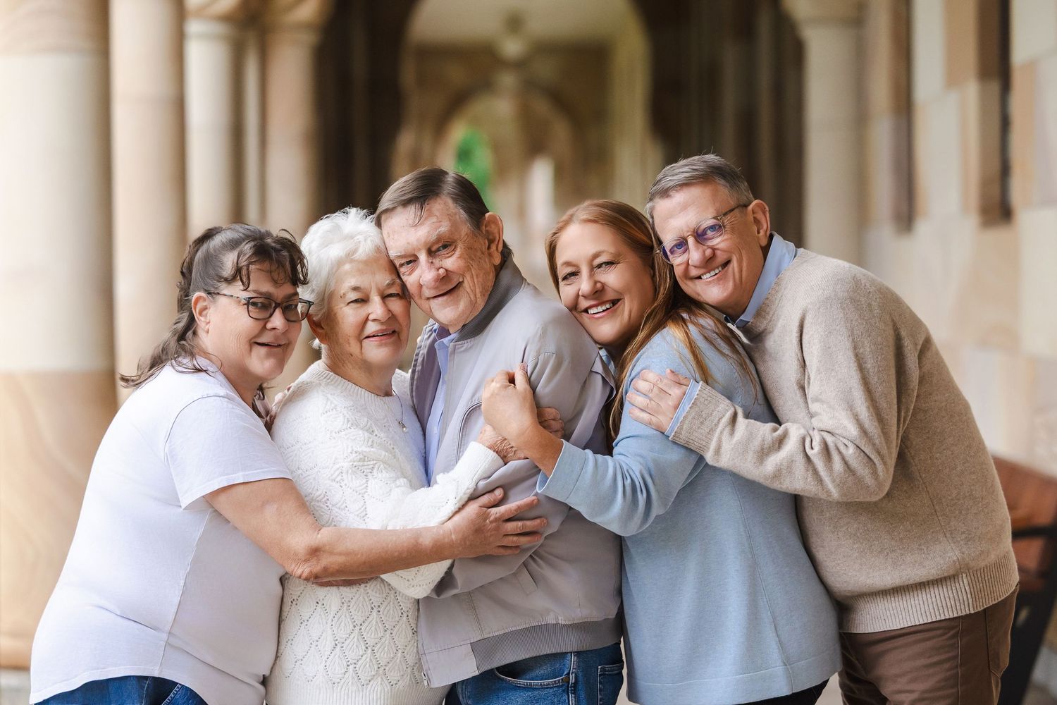 A cheerful multi-generational group hugs and laughs together while standing in an outdoor courtyard with columns.