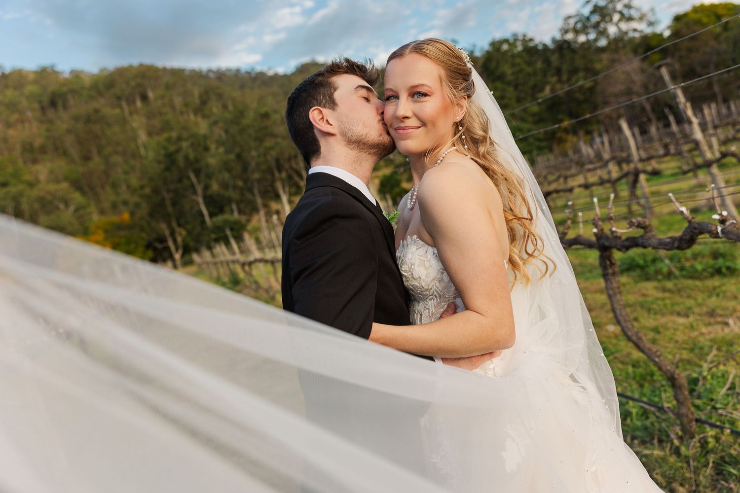 Groom kissing bride on cheek at vineyard wedding, her veil flowing in wind, lush green hills in background.